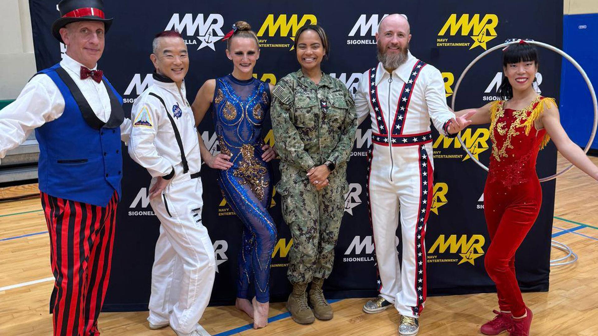 Freedom Circus performers pose with a Sailor in uniform in front of a Navy MWR Entertainment backdrop. The performers wear colorful costumes, including a ringmaster outfit, an astronaut-themed suit, and acrobatic attire.